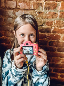 little girl holding digital camera in front of exposed brick wall