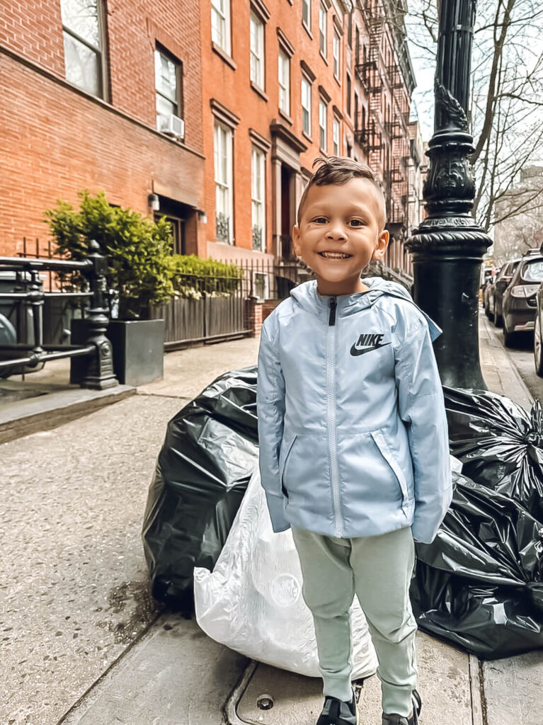 toddler boy in gray Nike jacket standing in front of black trash bags