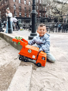 little boy with orange garbage truck in sand at park