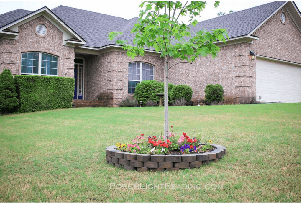 flowers around a tree with stone and mulch with brick house in the background