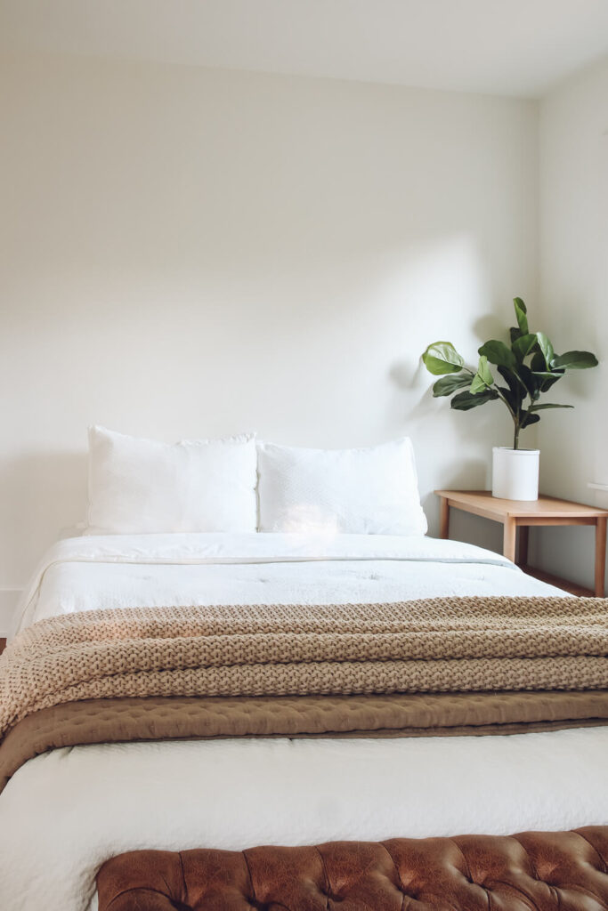 white walls in bedroom with white bedding and brown knit blanket with leather bench at the foot of the bed